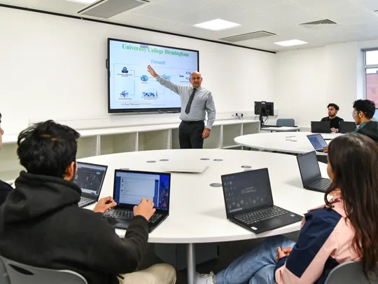 Male lecturer teaching a smaller group of students who are on laptops.